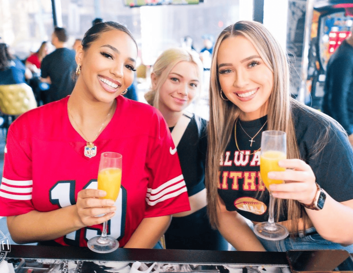 Three people sitting at a table, holding glasses of orange juice. Two are wearing sports-themed shirts, including a red NFL jersey and a black shirt with red and yellow text.